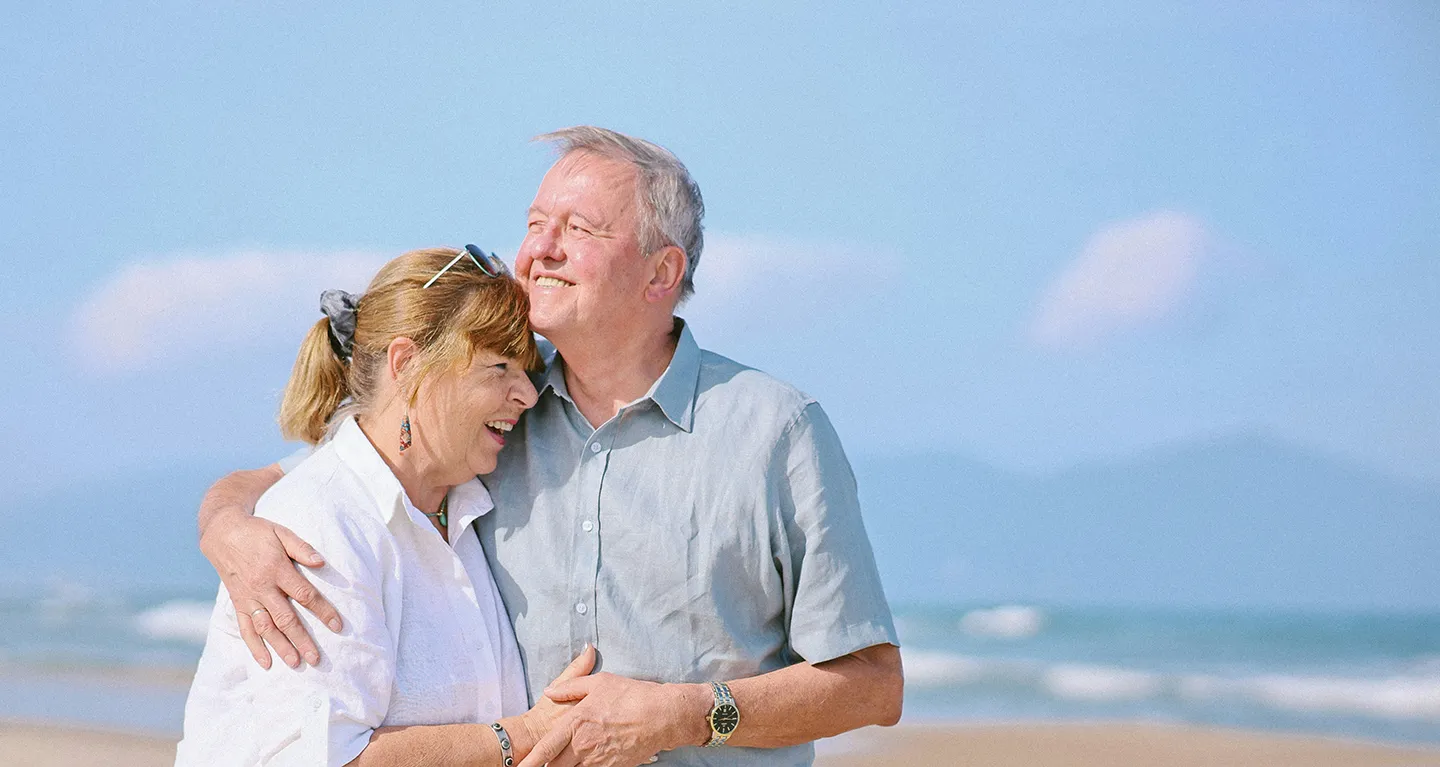 Retired couple on a sunny beach with arms around eachother in an embrace